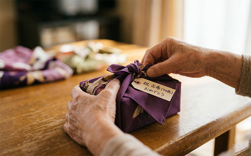 「祝 古希おめでとう」タグ付きの紫の風呂敷ギフトを開ける高齢者の手元