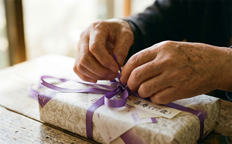 紫のリボンが結ばれた喜寿祝いの贈り物を開ける高齢者の手元