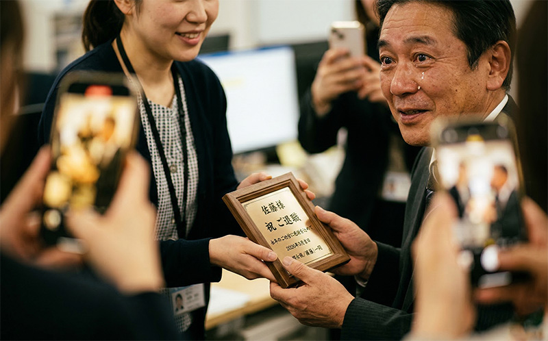 女性社員から額装ギフトを受け取り感動する退職者と撮影する同僚たち