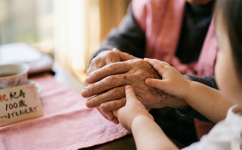 100歳の祖父の手と孫の小さな手が優しく重なるピンクの布の上