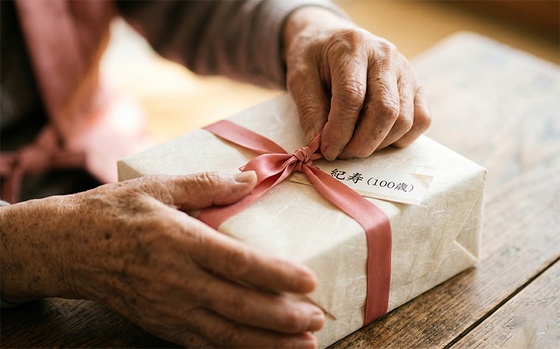 ピンクのリボンが結ばれた紀寿祝いのギフトを開ける100歳の手元