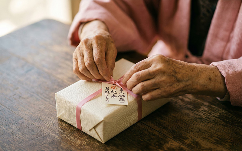 ピンクのリボンが結ばれた紀寿祝いのギフトを丁寧に開ける100歳の手元