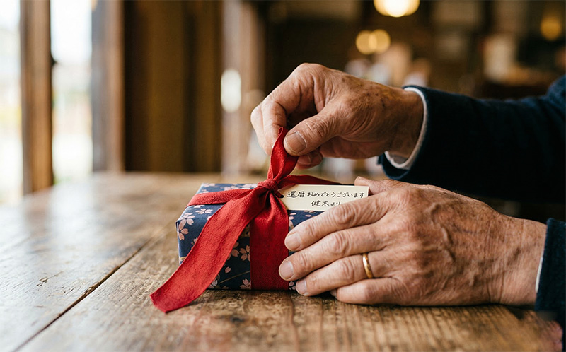還暦祝いの赤いリボンのギフトを開ける60歳男性の手元クローズアップ