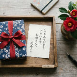 木製トレイに並ぶ赤いリボンのギフトと手書きメッセージカードと赤い花